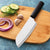 A Cook's Knife on a wood cutting board with garlic, cucumbers, and onions