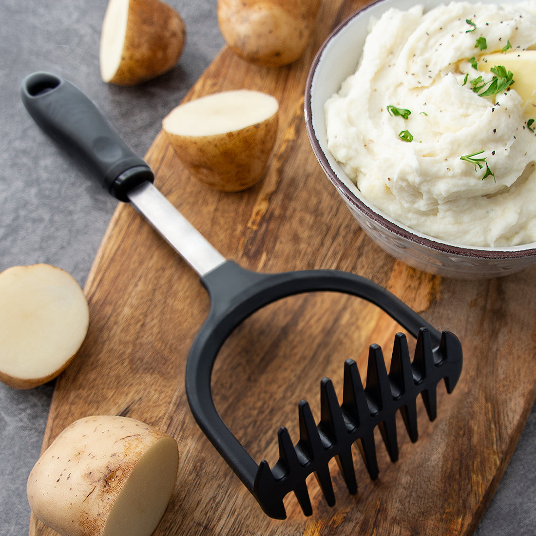 Rada Cutlery Potato Masher next to a bowl of mashed potatoes and whole potatoes on a cutting board.