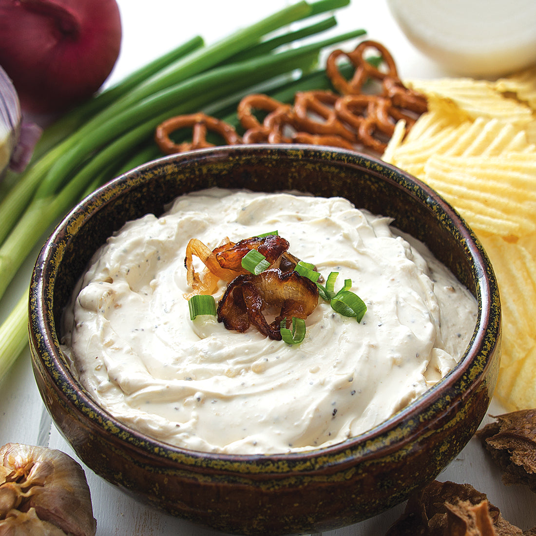 A bowl of Roasted Garlic & Onion Dip next to chips, pretzels. and green onions