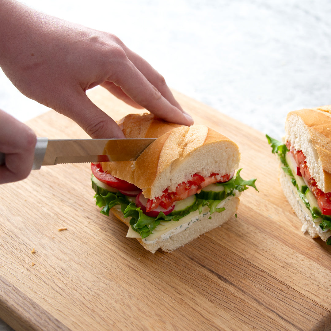 Bread Knife slicing through sandwich layered with tomatoes, radishes, lettuce, cucumber, cheese, and dill spread.