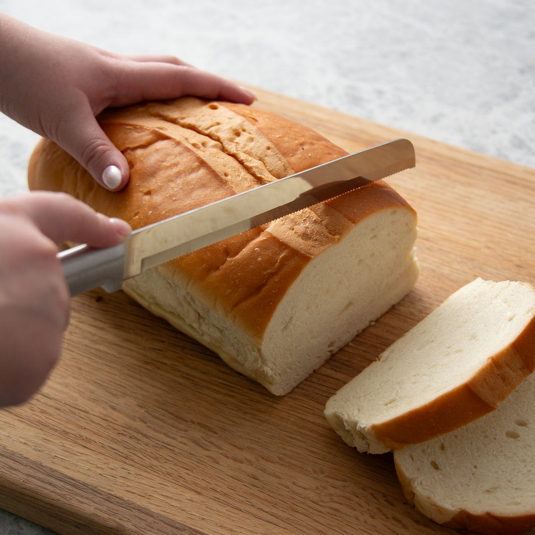 White bread being cut into slices with a serrated blade on a silver handle bread knife.