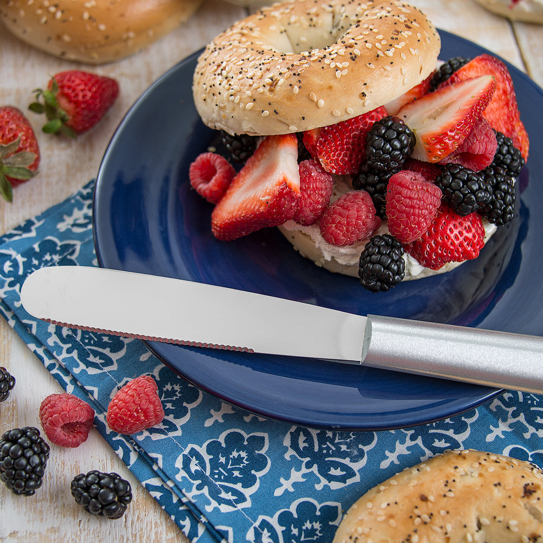 Rada Cutlery Super Spreader with a silver handle on a blue plate with a bagel, fruit, and napkin.