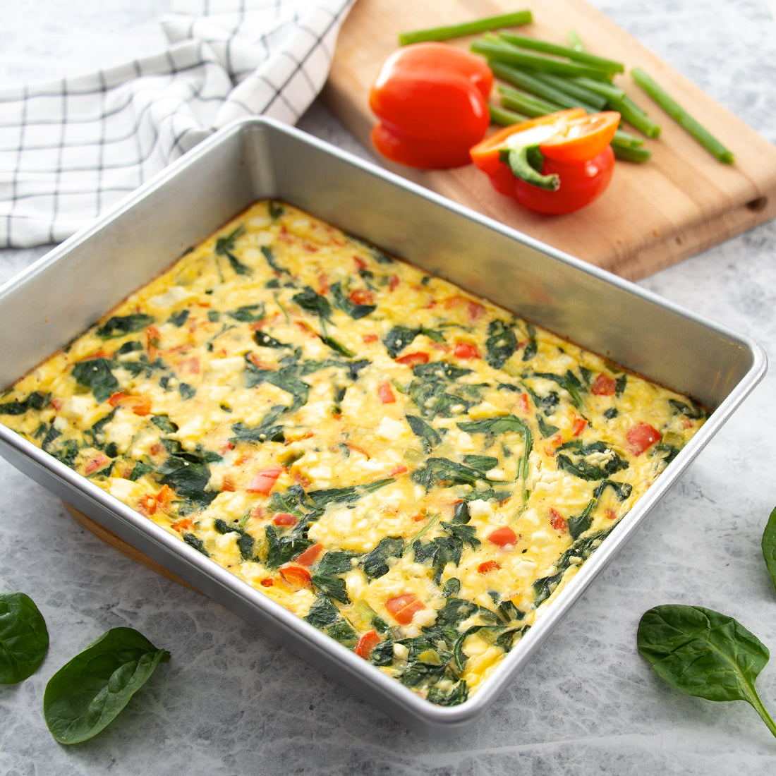 Rada Cutlery's Cake Pan used to make a baked spinach and tomato frittata, styled on a light gray surface with fresh spinach leaves, a cutting board with vegetables, and a checkered cloth.
