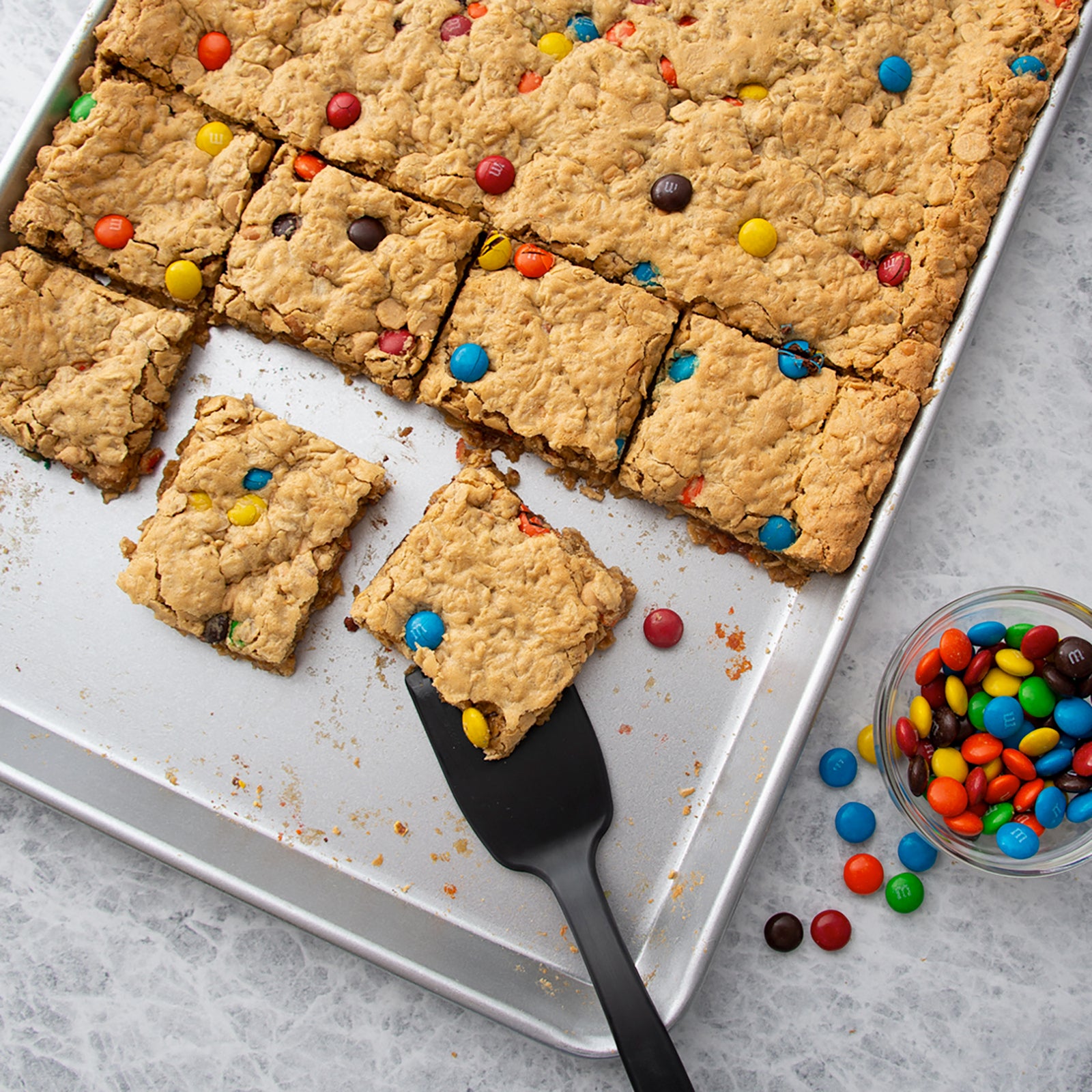 Rada Cutlery Half Sheet Pan with caramel oatmeal cookie bars topped with colorful M&M's candies, with a small glass bowl of candy-coated chocolate pieces on a light gray surface.