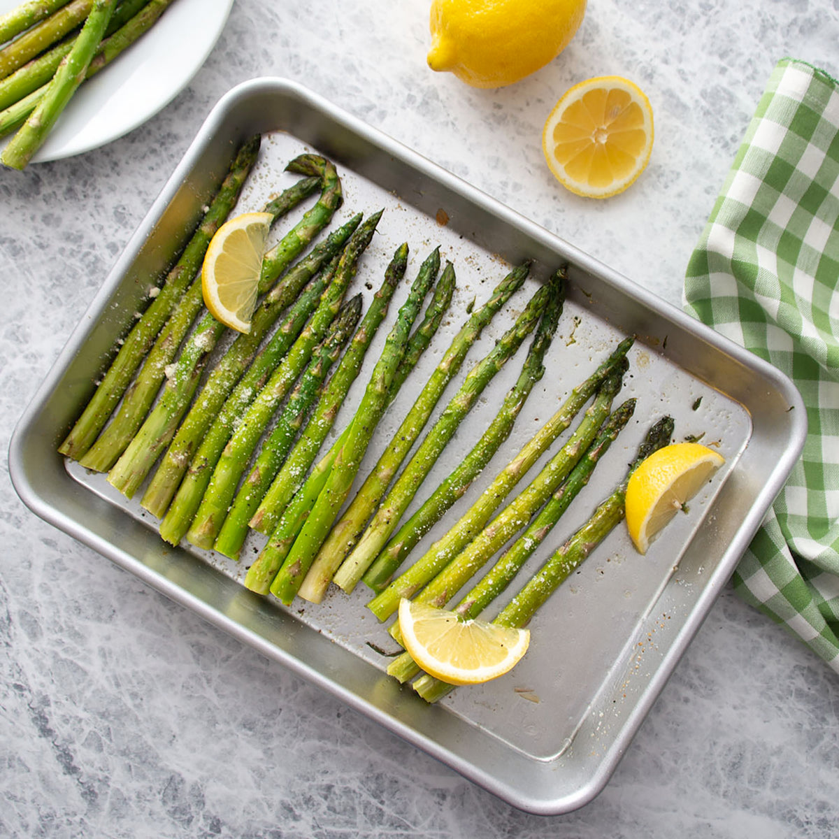Rada's Quarter Sheet Pan is used for roasted green asparagus spears with lemon slices, garnished with cheese, photographed from above on a light stone surface with lemons and a checkered cloth nearby.