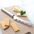 A Cheese Knife on a tan cutting board with assorted cheeses.