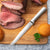 A silver handled Slicer on a cutting board with sliced roast beef and hamburger buns.