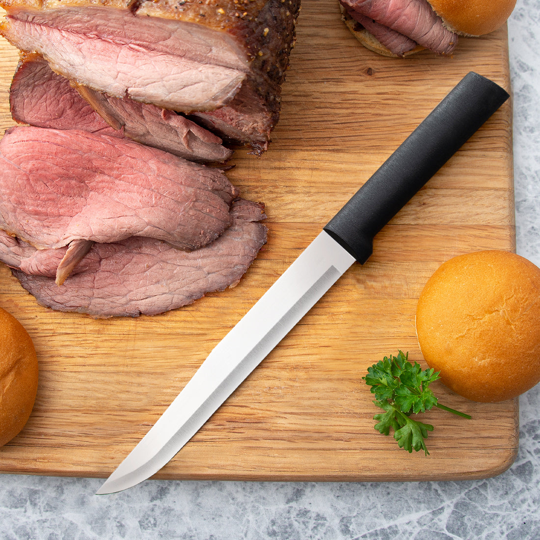 A silver handled Slicer on a cutting board with sliced roast beef and hamburger buns.