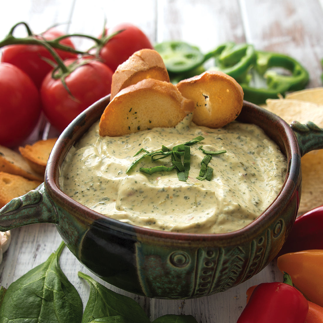 A bowl of Spinach Artichoke Dip with baguette slices next to tomatoes, peppers, chips, and spinach.