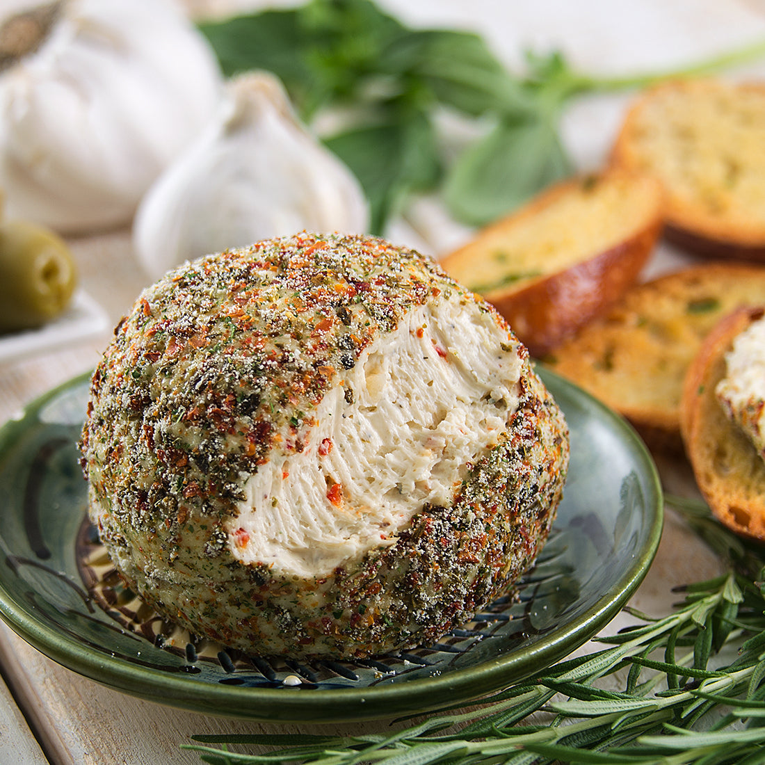 A Tuscan Herb Cheeseball on a green plate next to garlic, bread, olives, and fresh herbs.